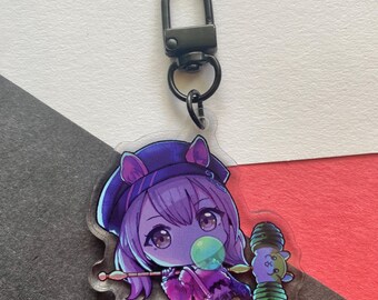 Op de afbeelding: Een sleutelhanger met een paars en blauw anime-personage met een zwarte clip. Het personage houdt een groene lolly en een klein pluche speelgoed.