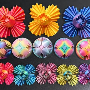 May include: An assortment of colorful, decorative objects in various shapes. The collection includes flower-like designs in red, yellow, and blue, along with circular pieces with intricate patterns and a rainbow color scheme. The objects are arranged on a dark surface.