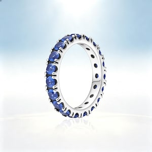 May include: A silver eternity band ring featuring a continuous row of round-cut blue sapphire gemstones set in a prong setting. The metal band has a polished finish and the stones are arranged around the entire circumference of the ring.