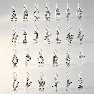 May include: A collection of twenty-six silver-toned metal letter charms, each featuring a small circular bail at the top and a series of embedded clear crystals along the front surface of every letter from A to Z, arranged in four rows.