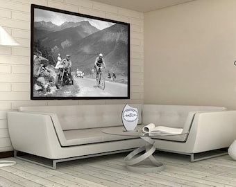 May include: A black and white photograph of cyclists racing up a mountain road. The photo is framed and hanging on a white wall in a living room. A white couch with a glass coffee table sits in front of the photo. A decorative metal sculpture sits on the table. A vase with a dried branch sits on the floor next to the couch.