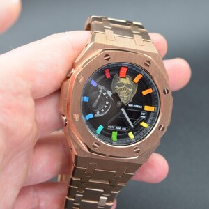 Puede incluir: Reloj de pulsera en tono oro rosa con esfera negra y diseño de calavera. El reloj tiene una caja rectangular y un brazalete de eslabones a juego. La esfera del reloj tiene detalles coloridos y el texto "WR 20BAR" y "MUTE ALM SIG".
