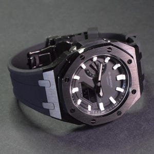 Op de afbeelding: Een zwart G-Shock horloge met een zwart rubberen bandje. De wijzerplaat van het horloge heeft een digitaal display en een witte analoge wijzerplaat met witte wijzers en markeringen. Het horloge heeft een zwarte metalen kast met een geborstelde afwerking.