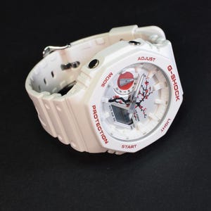 Peut inclure: Une montre G-Shock blanche avec un motif floral rouge et noir sur le cadran. La montre a un bracelet blanc et des boutons rouges. Le cadran de la montre porte les inscriptions "G-Shock", "Protection", "Mode", "Start", "Light" et "Adjust".