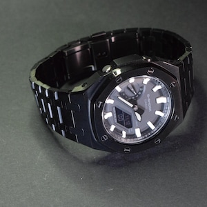 May include: A black G-Shock watch with a silver face and a black metal band. The watch has a digital display and a chronograph.