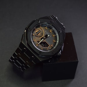 Op de afbeelding: Een zwart G-Shock horloge met een zwarte metalen band en een zwarte wijzerplaat. Het horloge heeft een digitaal display en een chronograaffunctie. Het horloge ligt op een zwart oppervlak.
