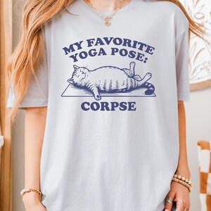 May include: A pale lilac t-shirt with navy blue text reading "MY FAVORITE YOGA POSE: CORPSE". The design includes a cat on a yoga mat. The t-shirt is a relaxed fit, suitable for casual wear.