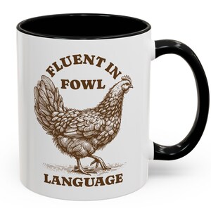 以下が含まれることがあります： 黒い内側とハンドルが付いた白いセラミックマグ。マグには、鶏の詳細な茶色のイラストと、茶色の文字で「FLUENT IN FOWL LANGUAGE」というテキストが描かれています。