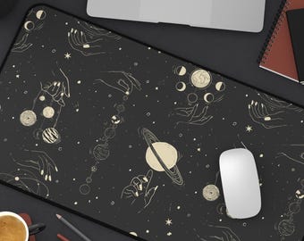 May include: A black rectangular desk mat with a cream celestial design of hands, planets, and stars. The mat has a black border and is shown with a laptop, mouse, coffee cup, and stationery.