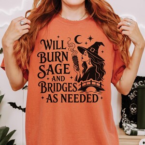May include: A burnt orange short-sleeve t-shirt featuring a black graphic print of a witch holding a smoking sage bundle above a bridge. The text reads: Will Burn Sage And Bridges As Needed, accompanied by small star and crescent moon icons.
