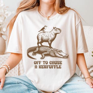 May include: Cream-colored t-shirt featuring a whimsical illustration of a capybara, bird, and alligator. The text "OFF TO CAUSE A KERFUFFLE" is printed below the animals in a brown font. The shirt is a relaxed fit.