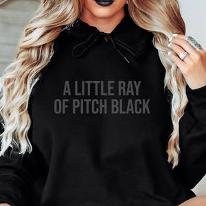 以下が含まれることがあります： グレーの文字で「A LITTLE RAY OF PITCH BLACK」と書かれた黒いフード付きスウェットシャツ。 スウェットシャツにはドローストリングフードとゆったりとしたフィット感があります。 画像は、人にスウェットシャツを着せている様子です。