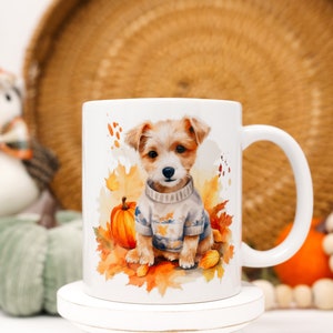 May include: White ceramic mug with a watercolor illustration of a small dog wearing a sweater and surrounded by autumn leaves. The dog is brown and white with a black nose and brown eyes. The mug has a handle on the right side.