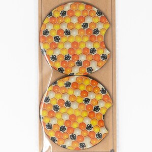 May include: Two round, wooden coasters with a honeycomb pattern in shades of yellow and orange. The honeycomb pattern is filled with small black bee silhouettes.