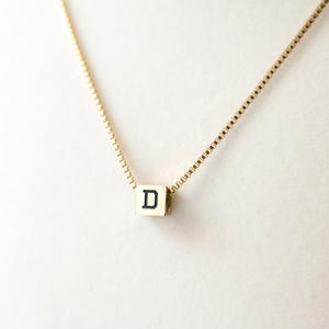 May include: A gold-toned box chain necklace featuring a small, square-shaped gold-toned pendant. The pendant is a cube with a black capital letter D printed on the front face. The chain is thin and has a square link pattern throughout its length.