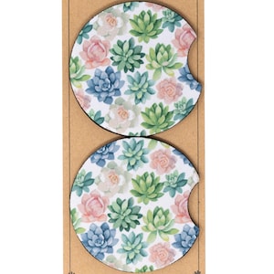 May include: Two round, white, cork-backed coasters with a floral pattern. The coasters feature a repeating pattern of green succulents and pink flowers.