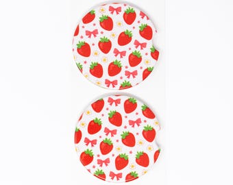 May include: Two white car coasters with a repeating pattern of red strawberries, pink bows, and small white flowers. Each coaster has a curved cutout for easy removal from the car cup holder.