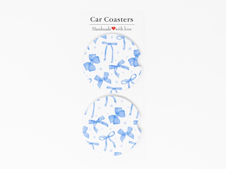 May include: Two white car coasters with a blue bow pattern. The coasters have a white background with blue bows and white stars. The text "Car Coasters" and "Handmade with love" is printed on a white card behind the coasters.