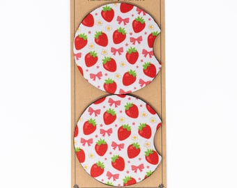 May include: Two circular car cup holder coasters featuring a repeating pattern of red strawberries, small white flowers, and pink bows on a white background. Each coaster has a small semi-circular notch cut out from the side for easy removal.