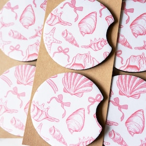 May include: A set of four circular car cup holder coasters featuring a white background with a repeating pattern of pink line-art seashells and small bows. Each coaster has a small semi-circular notch on one side and is packaged on brown cardstock.