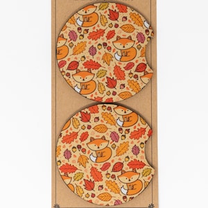 May include: Two round, brown coasters with a fall-themed design. The coasters feature orange foxes, brown acorns, and orange and yellow leaves on a beige background.