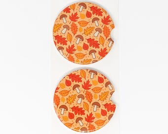 May include: Two round, orange and brown ceramic coasters with a pattern of autumn leaves and mushrooms. The coasters have a cut-out for a beverage holder.