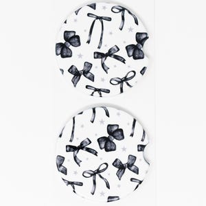 May include: Two white ceramic car coasters with a black and white bow and star pattern.