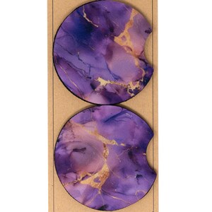 May include: Two purple and gold marble coasters with a cut-out for a cup. The coasters are round and have a marbled design with gold accents.