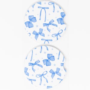 May include: Two white ceramic car coasters with a blue watercolor bow pattern. The coasters feature a repeating pattern of blue bows with white stars on a white background.