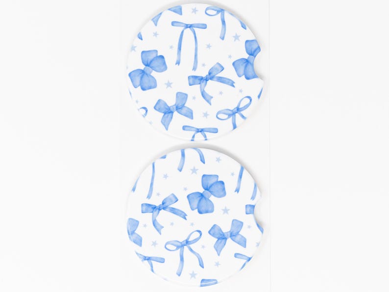 May include: Two white ceramic car coasters with a blue watercolor bow pattern. The coasters feature a repeating pattern of blue bows with white stars on a white background.