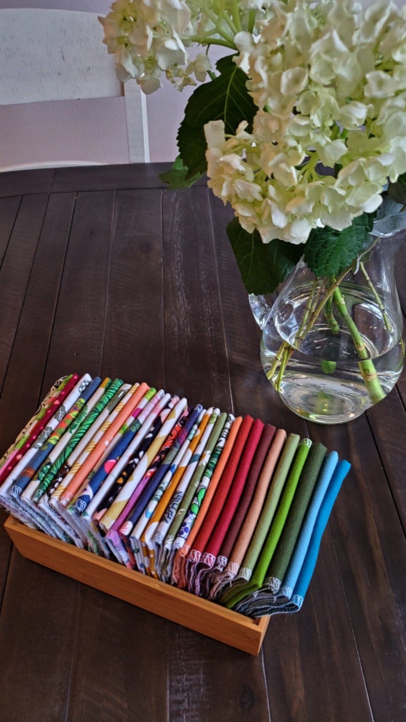 A table with a vase of hydrangea.  Next to it is a bamboo box with folded brightly colored and patterned napkins.  Each napkin edge is visible creating a rainbow effect.