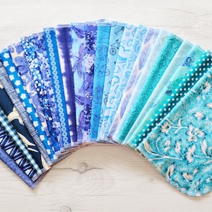 A fan of unpaper towels in an ombre of blue starting with navy, then royal blue, dusty blue, baby blue, then fading into teal and aqua blue.  Each pattern is different featuring a variety of vines, florals, nature, hearts, dots, stripes, bicycles.