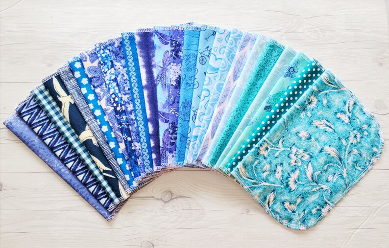 A fan of unpaper towels in an ombre of blue starting with navy, then royal blue, dusty blue, baby blue, then fading into teal and aqua blue.  Each pattern is different featuring a variety of vines, florals, nature, hearts, dots, stripes, bicycles.
