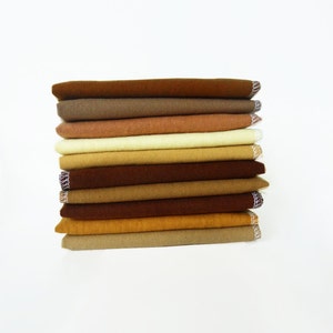 May include: A stack of folded fabric in various shades of brown and tan. The fabrics are neatly arranged, with visible edges and a textured appearance. The edges of the fabric have a decorative trim. The overall aesthetic is simple and clean.