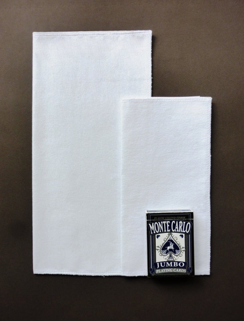 May include: Two white cotton handkerchiefs, one larger than the other, are laid out on a brown surface. A deck of blue and white Monte Carlo Jumbo Playing Cards is placed on top of the smaller handkerchief.