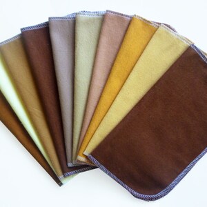 May include: A collection of ten rectangular cloth napkins in various shades of brown and yellow, fanned out to display the color variations. Each napkin has a rounded corner and a contrasting stitched edge.