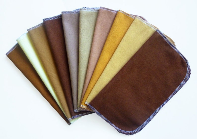 May include: A collection of ten rectangular cloth napkins in various shades of brown and yellow, fanned out to display the color variations. Each napkin has a rounded corner and a contrasting stitched edge.