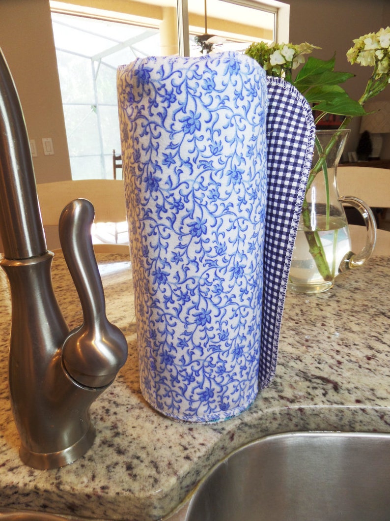 A roll of blue pattern unpaper towels next to a kitchen sink.  The main pattern is a tiny blue floral on white background.  The towel ready to be pulled off is a navy blue gingham.