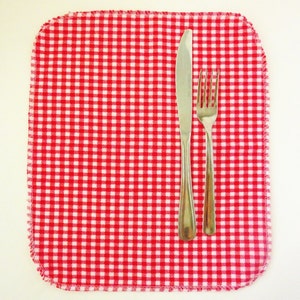 May include: A red and white checkered placemat with a stainless steel knife and fork. The placemat has rounded corners and a stitched edge. The silverware is placed vertically on the mat.