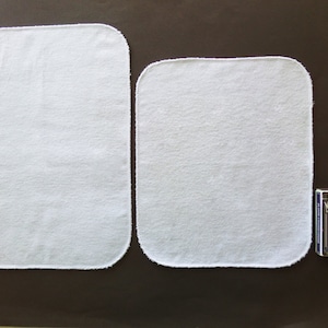 May include: Two white cotton cloths with a white stitched border. The cloths are rectangular and are lying flat on a brown surface. A deck of playing cards is visible in the background.