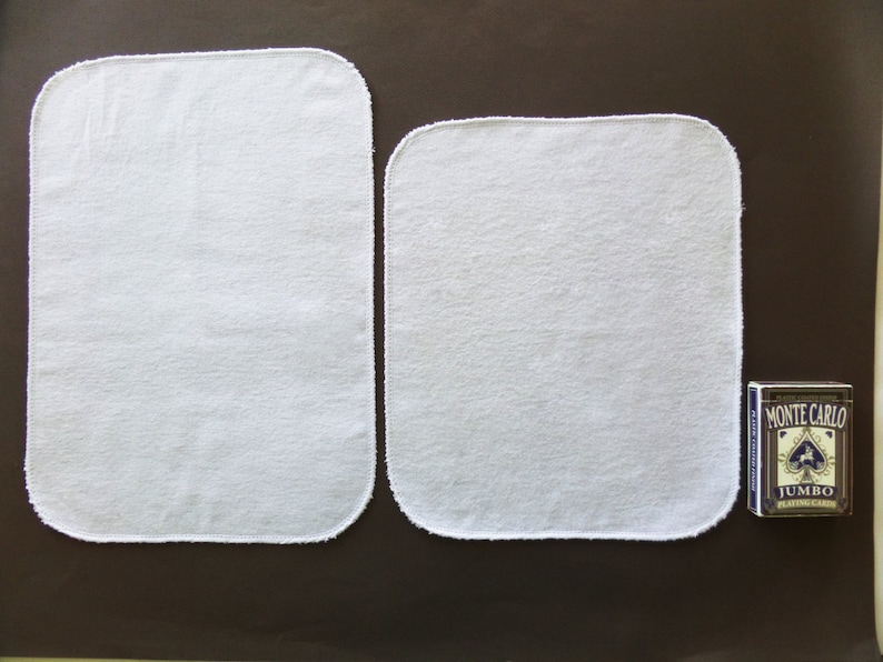 May include: Two white cotton cloths with a white stitched border. The cloths are rectangular and are lying flat on a brown surface. A deck of playing cards is visible in the background.