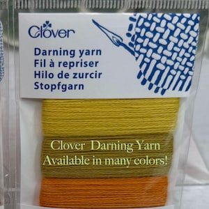 Op de afbeelding: Een verpakking Clover stopgaren in tinten geel en oranje. Het pakket bevat drie kleuren garen, elk 16 meter lang. De verpakking heeft de tekst "Clover Darning Yarn Available in many colors!"
