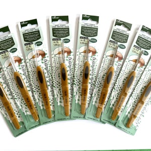 May include: A set of ten Clover Soft Touch Steel Crochet Hooks in various sizes, ranging from 1.75 mm to 0.50 mm. The hooks are gold and have ergonomic handles. The packaging is green and white with the text "Soft Touch Steel Crochet Hook" and "Ergonomic Crocheting".