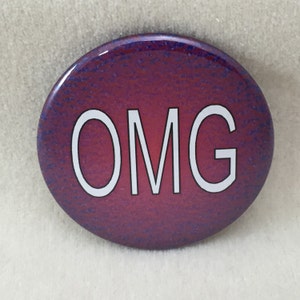 May include: A round button with a glossy finish. The button has a textured, dark red background with blue speckles. The white letters "OMG" are centered on the button.