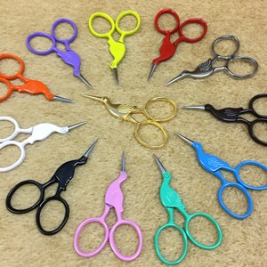 Può includere: Una collezione di dodici forbici da ricamo a forma di cicogna disposte in cerchio. Le forbici presentano vari colori solidi tra cui viola, giallo, rosso, arancione, bianco, nero, rosa, verde acqua, blu, marrone, oro e una finitura argento.