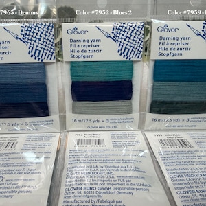 Op de afbeelding: Drie pakjes Clover stopgaren in blauwe, blauwgroene en denimtinten. Elk pakje bevat drie kleuren garen, met de tekst "Stopfgarn" en "16 m/17,5 yds" erop.