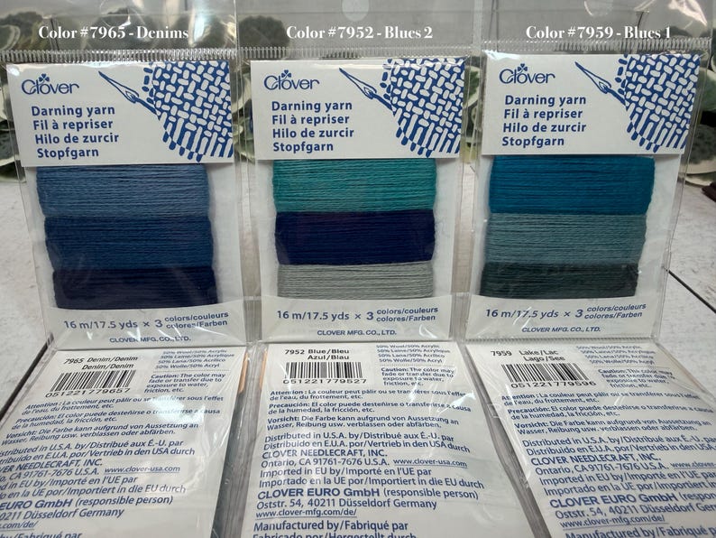 Op de afbeelding: Drie pakjes Clover stopgaren in blauwe, blauwgroene en denimtinten. Elk pakje bevat drie kleuren garen, met de tekst "Stopfgarn" en "16 m/17,5 yds" erop.