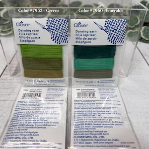 Op de afbeelding: Twee pakken Clover stopgaren in een doorzichtige plastic container. E&eacute;n pak is gelabeld "Greens" en de andere "Emeralds". Elk pak bevat drie tinten groen garen. De verpakking bevat de tekst "Stopfgaren".
