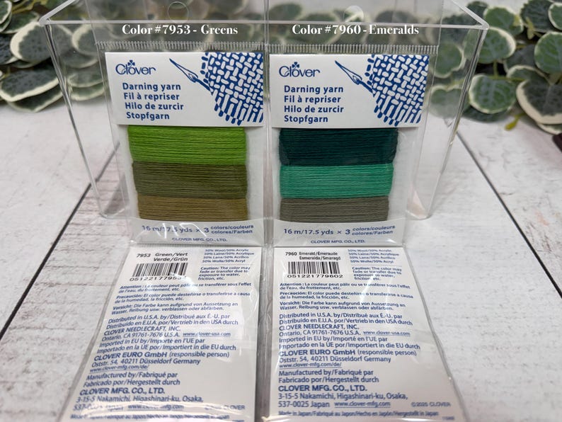 Op de afbeelding: Twee pakken Clover stopgaren in een doorzichtige plastic container. E&eacute;n pak is gelabeld "Greens" en de andere "Emeralds". Elk pak bevat drie tinten groen garen. De verpakking bevat de tekst "Stopfgaren".
