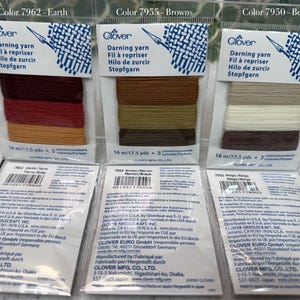 Op de afbeelding: Drie verpakkingen Clover stopgaren in de kleuren Aarde, Bruin en Beige. Elke verpakking bevat drie tinten garen, met 16 m per kleur. De verpakking toont het Clover-logo en productinformatie. Het garen is voor stoppen en repareren.